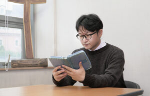 Rev Lee is seen seated at a table with a bible. He is wearing glasses and a dark sweater.