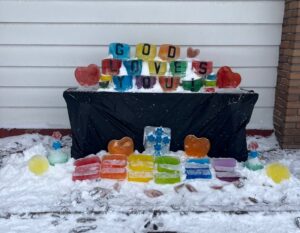 Beautiful rainbow coloured ice "glass" arranged on a table outside in the snow, with letters embedded in some of the blocks, spelling "God Loves You".
