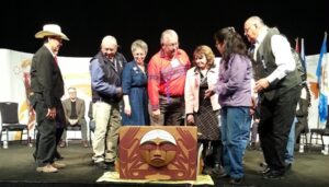 A group of eight people, Indigenous and non, stand around a beautiful bentwood box as items are placed in it at the Edmonton TRC national event. 