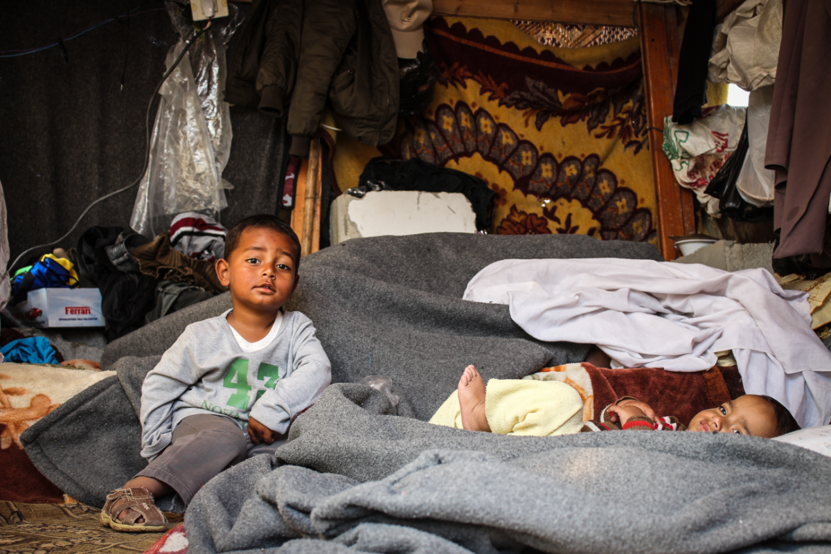 Two young children sitting under a makeshift tent made of clothing and blankets. Photo by Hosny Salah.