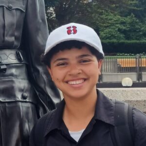 Photo of a Black person wearing a white ball cap, smiling into the camera. 