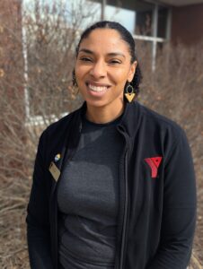 A Black woman standing outside, wearing a black top and earrings, smiling into the camera.