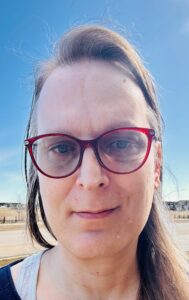 A white-presenting woman with glasses looking into the camera, blue sky in the background.