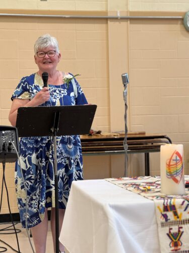 A smiling white woman with grey hair and blue and white dress speaking into a mic.